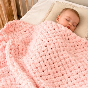 May include: A soft, pink, woven baby blanket covers a sleeping infant. The blanket has a thick, textured appearance and is draped over the baby, who is lying on a bed with pillows. The blanket is a light pastel pink color.
