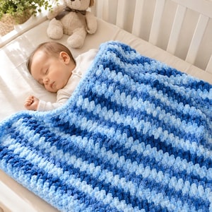 May include: A hand-crocheted baby blanket featuring a textured, chunky yarn design with alternating diagonal stripes in various shades of blue, ranging from light sky blue to deep navy, providing a soft and cosy appearance for a sleeping infant.