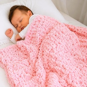 Op de afbeelding: Een slapende baby ligt onder een zachte, roze, dik gebreide deken. De deken is over de baby gedrapeerd, waardoor een gezellige en geruststellende scène ontstaat. De baby draagt een witte onesie en rust op een wit kussen.