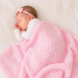 May include: A sleeping baby wearing a pink bow headband and a white long-sleeved shirt, covered with a soft, light pink crocheted blanket. The blanket has a delicate floral pattern.