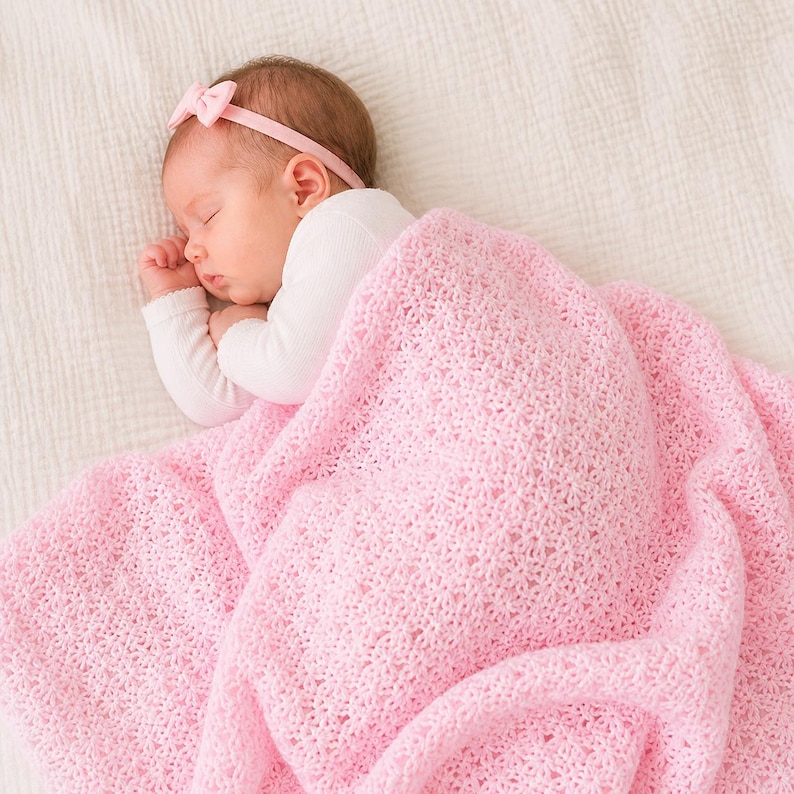 May include: A sleeping baby wearing a pink bow headband and a white long-sleeved shirt, covered with a soft, light pink crocheted blanket. The blanket has a delicate floral pattern.