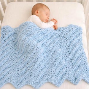 May include: A baby sleeps peacefully under a light blue crochet blanket with a scalloped edge. The blanket features a textured chevron design. The baby wears a white onesie and rests in a white cot. The blanket is approximately 100 cm x 100 cm.
