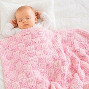 May include: A baby asleep under a pink and white chequered knitted blanket. The baby wears a white long-sleeved top and rests on a white pillow and sheets. The blanket has a soft, cosy design, perfect for a nursery.