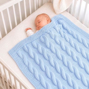 May include: A light blue knitted baby blanket featuring a prominent vertical cable knit pattern throughout the centre, bordered by a textured garter stitch edge. The soft, woven fabric is draped over a sleeping infant in a white cot.