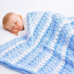 Può includere: Un bambino che dorme è rannicchiato sotto una morbida coperta all'uncinetto. La coperta presenta un motivo bianco e azzurro, creando una scena accogliente e confortante. Il bambino indossa una maglietta bianca a maniche lunghe.
