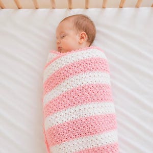 May include: A sleeping baby wrapped in a pink and white striped crochet blanket. The blanket is in a crib with wooden rails. The baby is swaddled in the blanket, with only the head visible. The blanket is approximately 30 inches long.