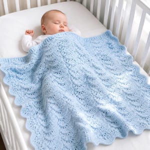 May include: A sleeping baby lies in a white cot, covered by a light blue, scalloped-edge knitted blanket. The blanket features a delicate, openwork design. The baby wears a white long-sleeved onesie.