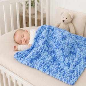 May include: A baby wearing a white long-sleeved shirt lies on a beige mattress, covered by a chunky, hand-knitted blanket featuring a variegated pattern of light blue, medium blue, and dark blue yarn. A light brown plush teddy bear sits nearby.