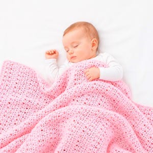 May include: A sleeping baby is nestled under a soft, pink, crocheted blanket. The blanket has a delicate, openwork pattern. The baby is wearing a white long-sleeved shirt. The background is a plain white surface.
