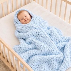 May include: A baby wrapped in a light blue, chunky knit blanket inside a wooden crib. The blanket is soft and fluffy, with a hood that covers the baby's head. The baby is looking at the camera and smiling.