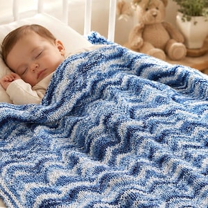 May include: A hand-knitted baby blanket featuring a wavy chevron pattern in various shades of blue and white. The blanket is textured with a soft yarn construction and displays a repeating ripple design that covers the entire surface of the fabric.