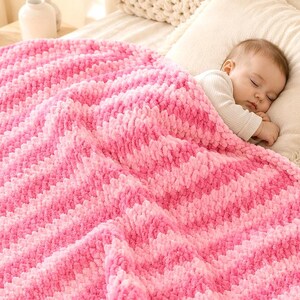 Peut inclure: Une couverture en tricot épais et doux avec un motif à rayures diagonales alternant des tons de rose clair et de rose vif. La couverture présente une texture en boucles de laine pelucheuses et recouvre un nourrisson endormi en haut blanc.
