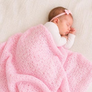 May include: A sleeping baby wrapped in a soft, light pink crocheted blanket. The baby wears a white long-sleeved shirt and a pink headband with a bow. The blanket has a delicate, openwork pattern.