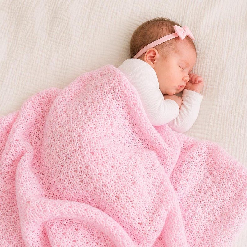 May include: A sleeping baby wrapped in a soft, light pink crocheted blanket. The baby wears a white long-sleeved shirt and a pink headband with a bow. The blanket has a delicate, openwork pattern.