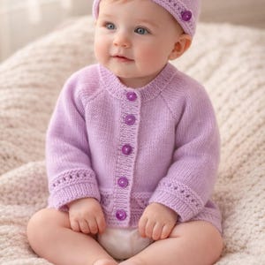 May include: A baby wearing a matching lavender knit cardigan and beanie hat. The cardigan has purple buttons down the front and decorative stitching on the cuffs. The baby is sitting on a cream-colored knitted blanket.