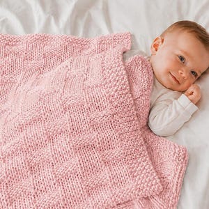 May include: A soft, light pink knitted baby blanket featuring a textured basketweave pattern. The blanket is constructed with thick yarn and has a distinct border, showcasing a chunky knit design that provides a tactile, raised surface appearance.