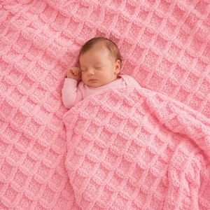 May include: A soft, light pink knitted blanket featuring a raised waffle-like grid texture. The blanket is draped over a sleeping infant who is wearing a long-sleeved light pink shirt, with the textured fabric covering part of the infant's body.