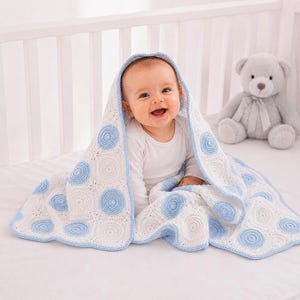 May include: A white and light blue crocheted baby blanket featuring a repeating pattern of circular motifs. The blanket is draped over a baby wearing a white long-sleeved shirt, with a gray plush teddy bear sitting nearby on a white surface.