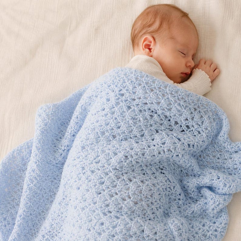 May include: A sleeping baby is wrapped in a light blue crocheted blanket. The blanket has a textured, open weave pattern. The baby is wearing a white long-sleeved shirt.