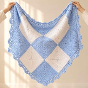 Könnte beinhalten: Eine quadratische Häkeldecke mit einem Schachbrettmuster aus hellblauen und weißen rautenförmigen Motiven. Die Decke ist mit einem dekorativen, wellenförmigen Rand versehen, der das gesamte Stück in hellblauem Garn einfasst.