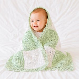 May include: A baby wrapped in a handmade crochet blanket featuring a checkerboard pattern of alternating sage green and white square motifs. The blanket is finished with a delicate scalloped lace border along the edges, showcasing intricate yarn work.