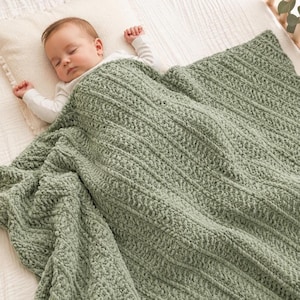 Könnte beinhalten: Ein Baby in einem weißen Langarmshirt liegt auf einem weißen Kissen, teilweise bedeckt von einer salbeigrünen, grob gestrickten Decke. Die Decke weist ein strukturiertes Rippenmuster mit dicken, weichen Garnschlaufen auf, die warm wirken.