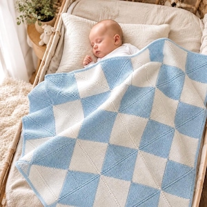 May include: A sleeping baby is nestled in a wooden bassinet, covered by a soft, knitted blanket. The blanket features a geometric pattern of alternating light blue and white squares. The baby is wearing a white onesie.