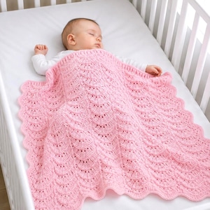 May include: A baby asleep in a white cot, covered by a pink, scalloped-edge knitted blanket. The blanket features a detailed, openwork pattern. The baby is wearing a white long-sleeved top.