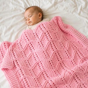 May include: A soft, light pink knitted baby blanket featuring a repeating diamond pattern with openwork detailing. The blanket is crafted from thick yarn and covers the upper body of a sleeping infant who is wearing a plain, light-coloured shirt.