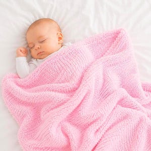 Puede incluir: Un bebé dormido envuelto en una suave manta rosa. El bebé lleva un body blanco y está acostado sobre una sábana blanca. La manta tiene un aspecto texturizado y afelpado, creando una escena acogedora y reconfortante.