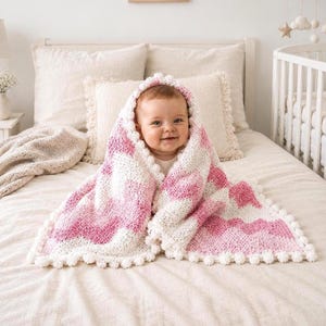 Könnte beinhalten: Eine handgehäkelte Babydecke mit einem wellenförmigen Zickzackmuster in abwechselnden Streifen aus zartem Rosa und Weiß. Die Decke ist mit einer dekorativen Bordüre aus kleinen, runden weißen Bommeln entlang der gesamten Außenkante versehen.