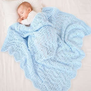 Può includere: Un bambino che dorme è coperto da una coperta azzurra a trama aperta. La coperta ha un bordo smerlato e un delicato motivo a pizzo. Il bambino indossa un body bianco e riposa su un cuscino bianco.
