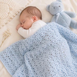May include: A sleeping baby is nestled under a light blue crocheted blanket. A blue teddy bear with a ribbon sits nearby. The blanket has a delicate openwork pattern. The baby is wearing a white onesie. A sprig of dried flowers is on the left.