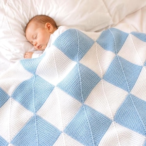 Puede incluir: Un bebé dormido está cubierto con una manta de punto azul y blanco. La manta presenta un patrón geométrico de diamantes. El bebé está acostado sobre una sábana blanca.