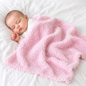 May include: A baby sleeps peacefully under a soft, pink blanket on a white bed. The blanket is a light pastel pink and has a textured appearance. The baby is wearing a white onesie, creating a serene scene.