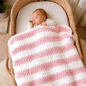 Puede incluir: Un bebé durmiendo en una cuna de mimbre, cubierto con una manta suave a rayas. La manta presenta franjas anchas alternas de color blanco y rosa claro. El bebé lleva un body blanco.