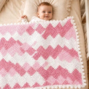 May include: A handmade crochet baby blanket featuring a chevron pattern in alternating stripes of white, light pink, and dark pink yarn. The rectangular blanket is finished with a white pom-pom trim along the edges and has a textured yarn finish.