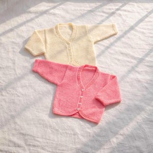 Puede incluir: Dos cárdigans de bebé tejidos. Uno es de color crema suave y el otro rosa claro. Ambos tienen cuello en V y cierre de botones. Los cárdigans están hechos de un hilo suave y texturizado.