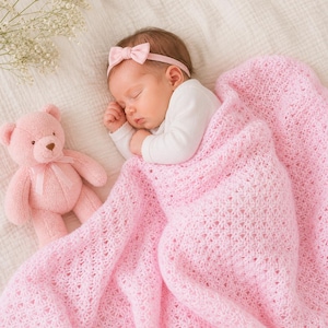 May include: A sleeping baby wrapped in a pink, open-weave blanket, wearing a pink bow headband and a white onesie. A pink teddy bear with a matching bow sits nearby. Soft, neutral background with delicate white flowers.