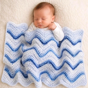 May include: A sleeping baby is nestled under a blue and white chevron-patterned crochet blanket. The blanket's design features varying shades of blue and white, creating a textured, handmade appearance. The baby is wearing a white onesie.