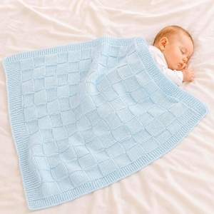 May include: A light blue knitted baby blanket with a woven pattern. The blanket is rectangular and has a textured border. A sleeping baby is partially visible, resting on a white surface. The blanket is soft and cozy.