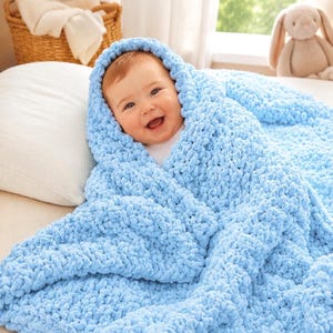 May include: A baby wrapped in a soft, light blue, chunky knit blanket. The blanket is draped over the baby's head, creating a cozy, hooded effect. The baby is smiling. A plush bunny is in the background.