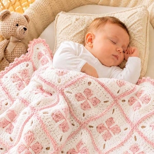 Può includere: Un bambino che dorme è rannicchiato in una culla, coperto da una coperta all'uncinetto bianca e rosa fatta a mano. Un orsacchiotto beige con un fiocco è vicino. La coperta presenta un motivo floreale.