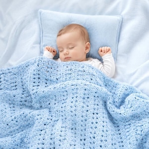May include: A sleeping baby lies on a light blue pillow and is covered with a light blue crocheted blanket. The baby is wearing a white long-sleeved shirt. The blanket has a delicate, openwork pattern.