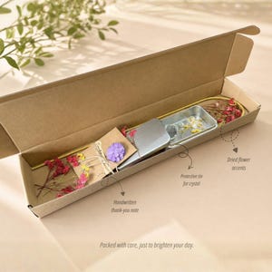 May include: A gift box with a handwritten thank-you note, a protective tin, and dried flower accents. The box is open, revealing the contents, including a small purple flower and a metal tin. The text on the box says, "Packed with care, just to brighten your day."