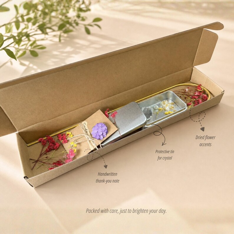 May include: A gift box with a handwritten thank-you note, a protective tin, and dried flower accents. The box is open, revealing the contents, including a small purple flower and a metal tin. The text on the box says, "Packed with care, just to brighten your day."