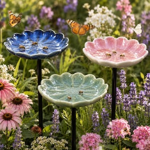 Peut inclure: Trois bassins d'eau en céramique en forme de fleur, de couleur bleue, rose et vert clair, montés sur de fines tiges métalliques noires. Les bassins sont remplis d'eau et attirent des abeilles, avec des papillons orange et blancs à proximité.