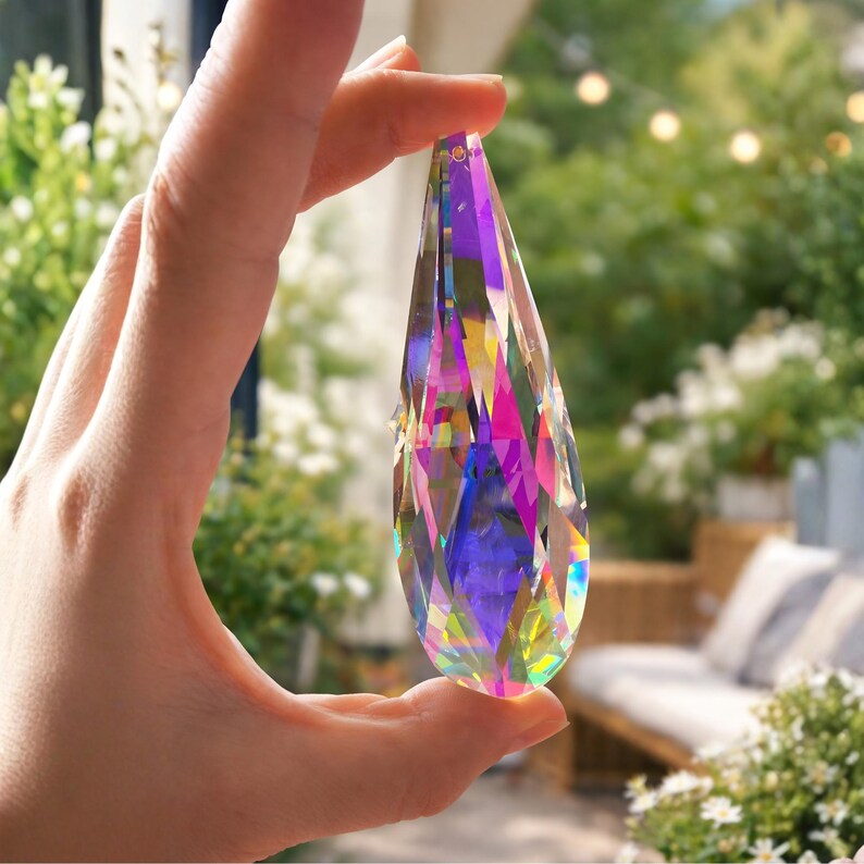 May include: A clear, teardrop-shaped crystal prism with multiple facets, reflecting a spectrum of colors including pink, purple, yellow, and blue. The prism is held up against a blurred background of greenery and outdoor seating.