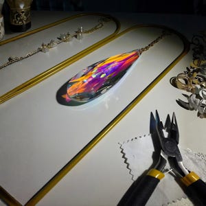 May include: A teardrop-shaped crystal pendant with iridescent colors of orange, purple, and yellow hangs from a gold chain. The pendant is displayed on a white surface with gold-colored metal frames. Jewelry-making tools are also visible.