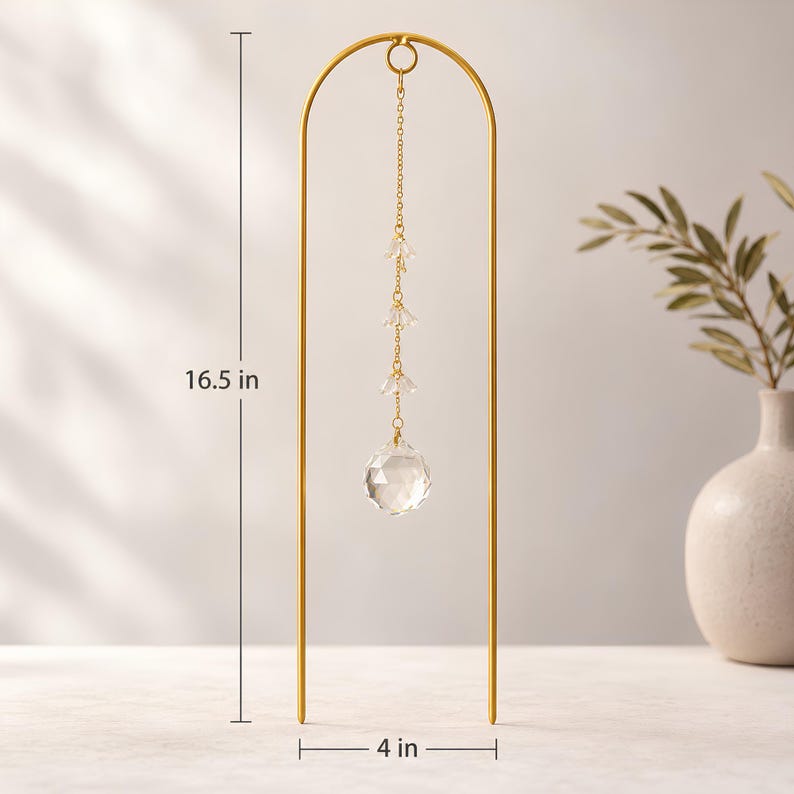 May include: A gold-colored metal arch with a hanging crystal ornament. The ornament is suspended from a chain and features a faceted crystal ball and smaller crystal accents. The arch is 16.5 inches tall and 4 inches wide.
